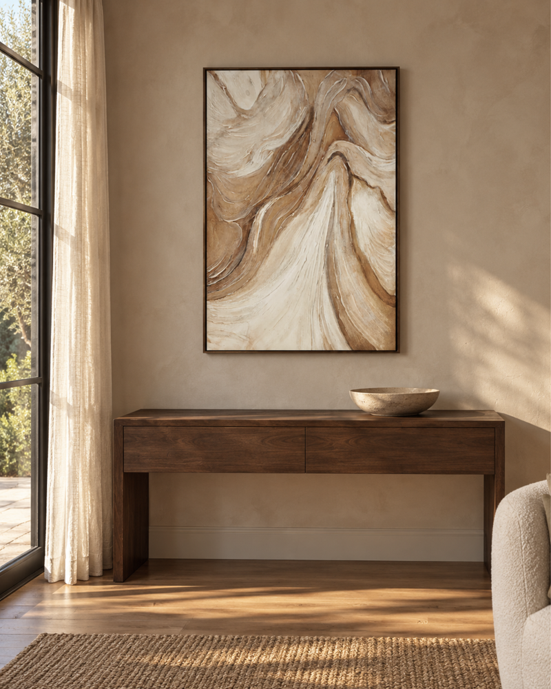 A framed abstract wall art print with flowing beige, cream, and brown marbled swirls hangs above a dark wood console table in a warm, sunlit room with a window and sheer curtain.