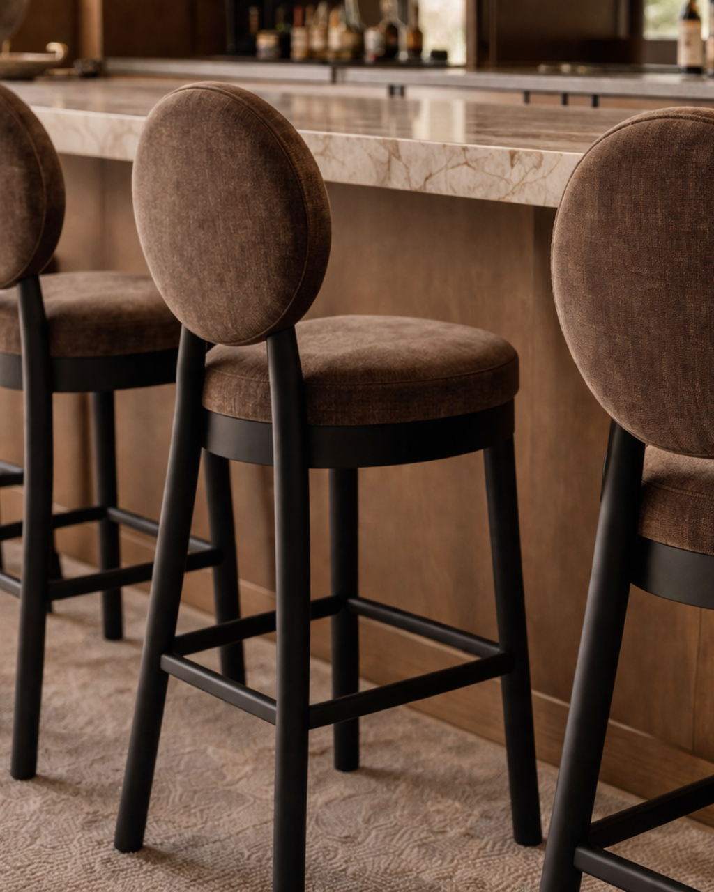 Set of contemporary bar stools with round brown upholstered seats and curved backrests, featuring black wooden legs, styled beside a marble kitchen island