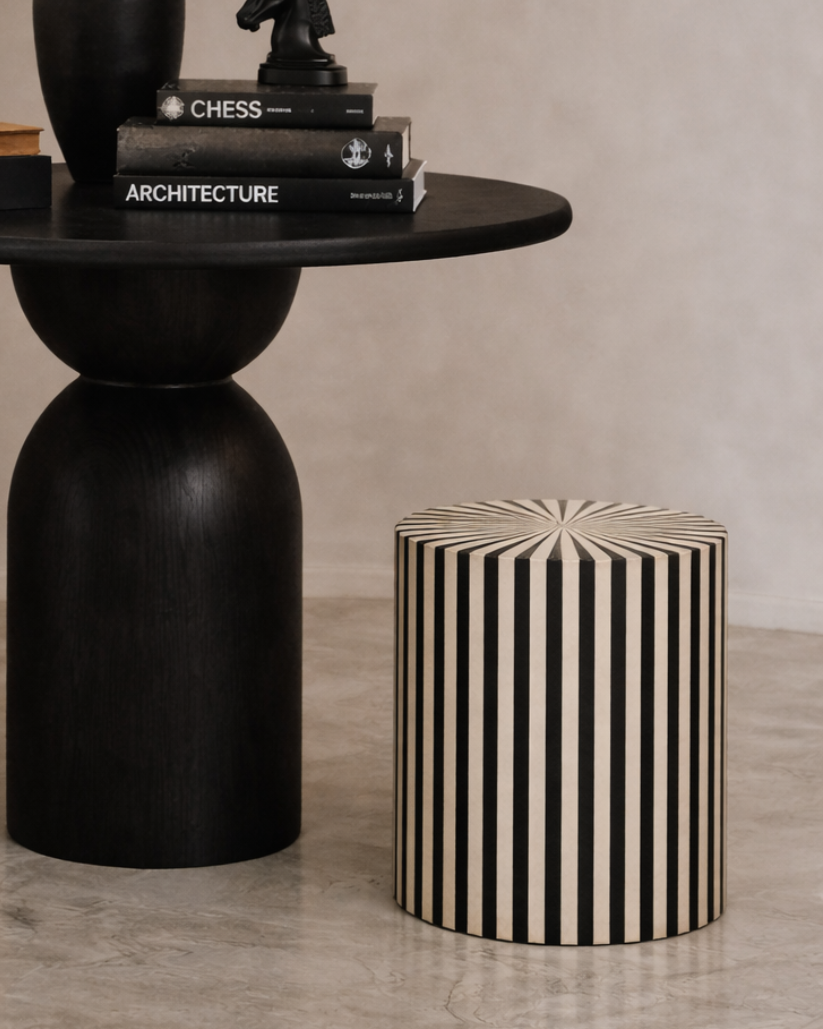 Contemporary interior scene featuring a graphic monochrome accent stool with vertical stripes next to a dark wood round table with curved pedestal base, styled with books and ornaments.
