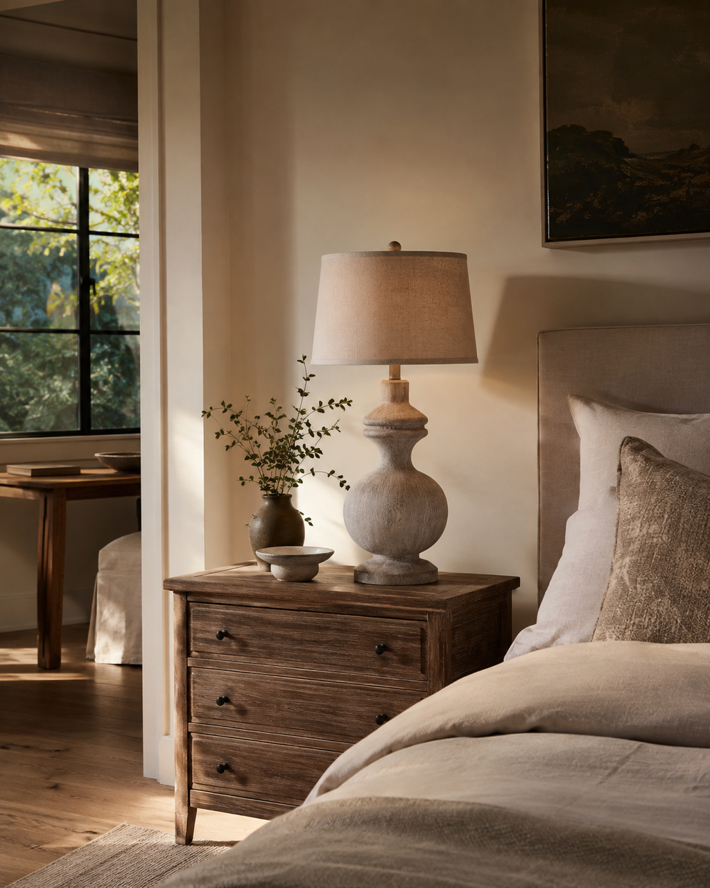 A rustic ceramic table lamp with a beige fabric drum shade and textured stone-like base, styled on a dark wood nightstand in a warm, neutral-toned bedroom.