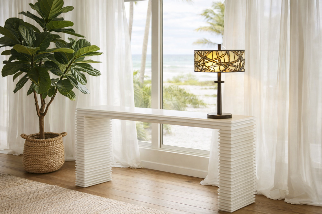 White textured console table with a lamp and plant in a bright room with large windows.
