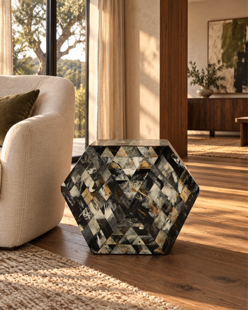 A hexagonal side table with a geometric mosaic shell pattern in black, white, gray, and gold, styled in a warm, modern living room next to a beige armchair.