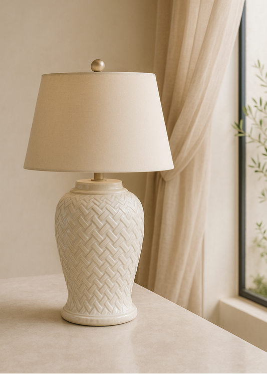 White textured table lamp with beige lampshade on a surface near a window with curtains.