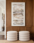 Two round cream upholstered ottomans with layered cushion-style bases sit side by side against a rich dark wood panelled wall. Above them hangs a large framed abstract canvas in warm neutral tones of white, beige, taupe, and charcoal. A woven natural rug lies beneath, creating a calm, sophisticated interior scene.