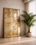 Large antiqued gold floor mirror leaning against a microcement wall beside a tall palm in a stone planter, with sheer white curtains and warm natural light.