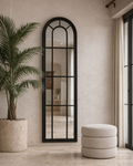 Decorative mirror with black frame on a wall next to a potted plant and round ottoman.