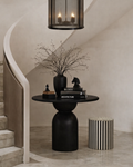 A black and ivory striped cylindrical stool sits on a polished stone floor beside a sculptural black pedestal side table topped with stacked books and decorative objects.