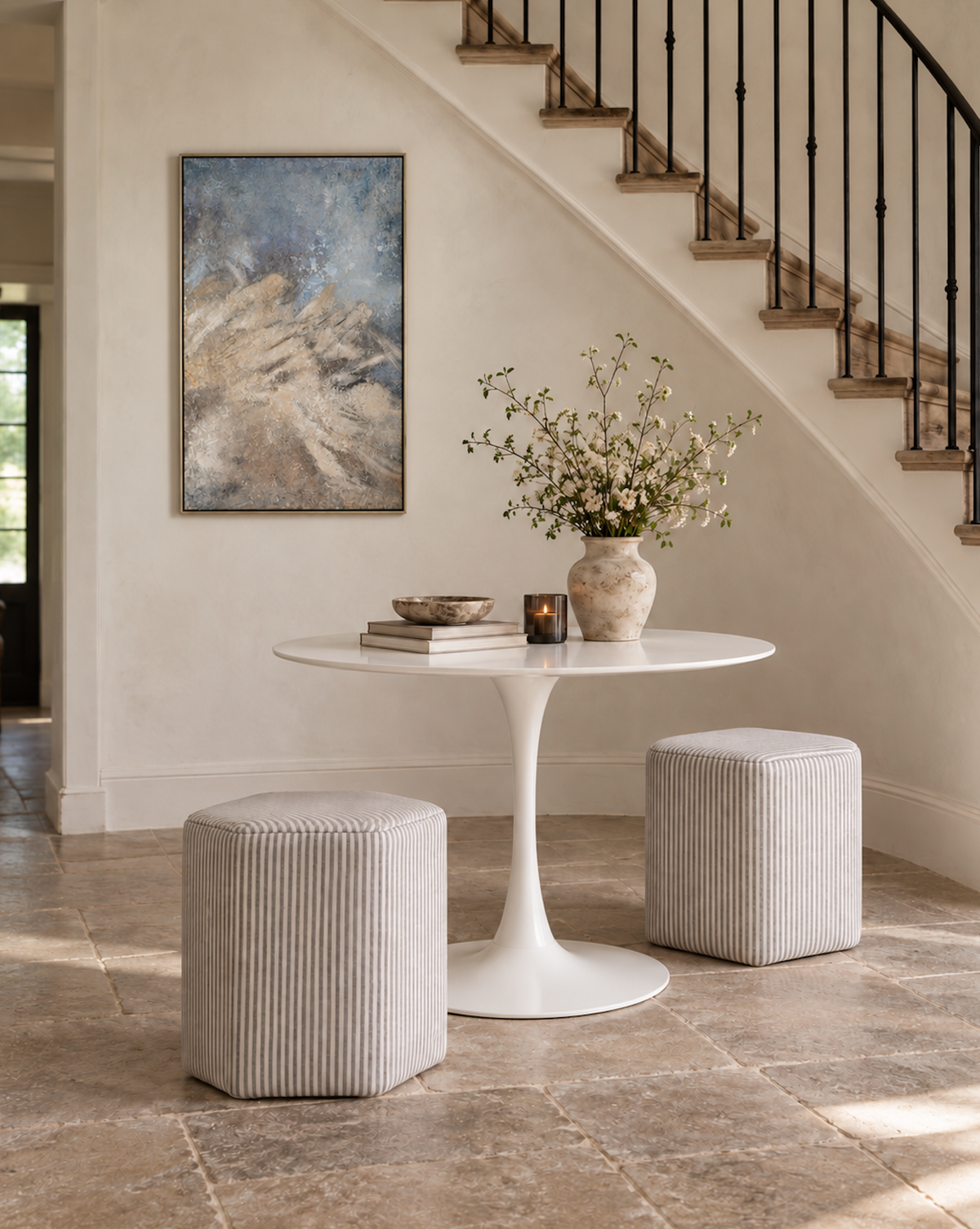 A styled interior scene with a round white pedestal table, two ribbed light beige poufs, a ceramic vase with branches, books, and a candle on the tabletop, plus a staircase and framed abstract wall art in the background.