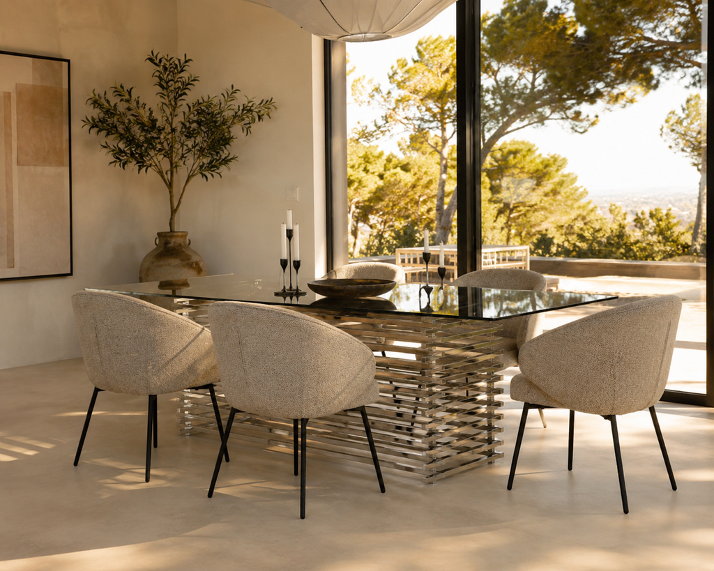 A modern dining set featuring a rectangular glass-top table with a sculptural metallic base and four upholstered dining chairs in a light beige textured fabric, set in a bright, sunlit room with large windows overlooking a garden.
