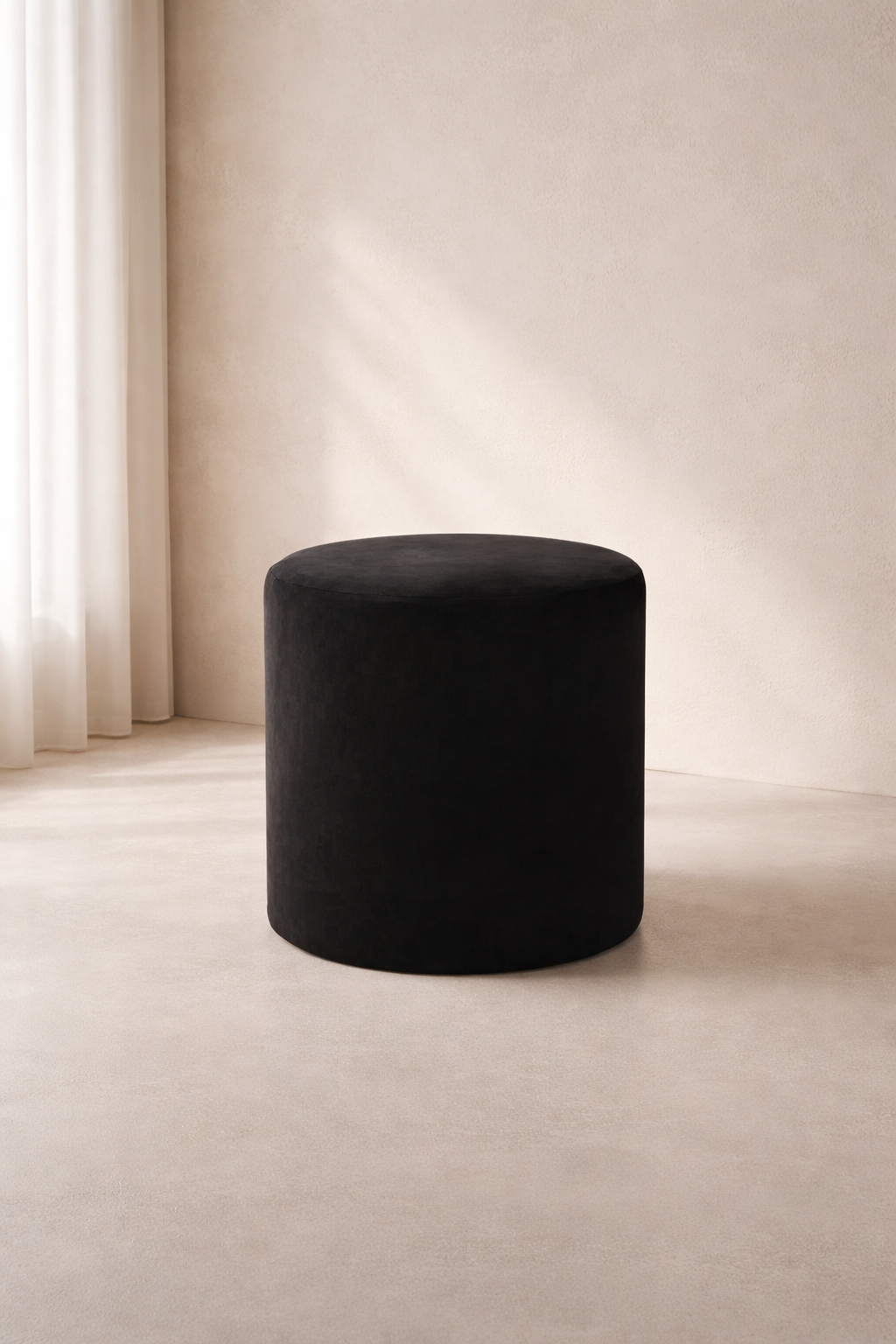 Black velvet cylindrical stool (38 cm high, 35 cm diameter) photographed in a minimalist studio with a warm neutral plaster wall and pale microcement floor, softly lit by natural daylight through sheer curtains.
